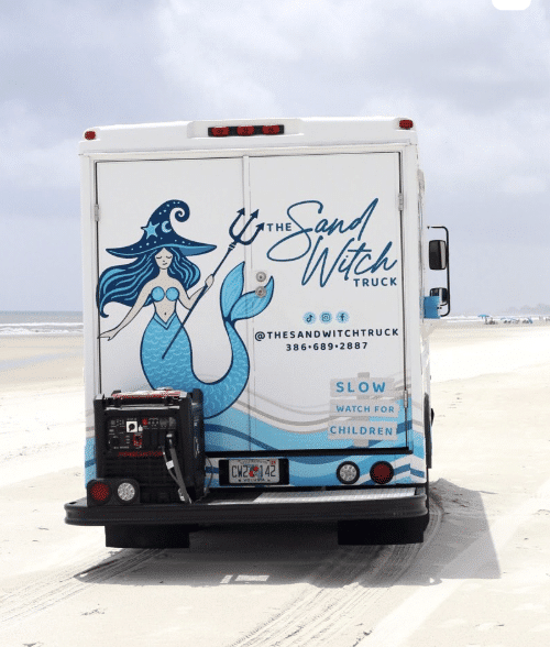 The SandWitch Truck on the beach and catering, rear view. A mermaid with a trident adorns the truck, promising beachside sandwiches. Watch for children! Slow.
