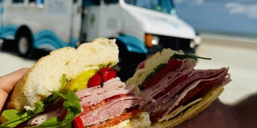 Beachside bliss! Two halves of a delicious SandWitch sandwich are held up against a backdrop of white sand, blue sky, and The SandWitch Truck. Lunch with a view!